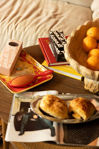 The red Detangling Brush on a tray with Greedy books, oranges and pastries